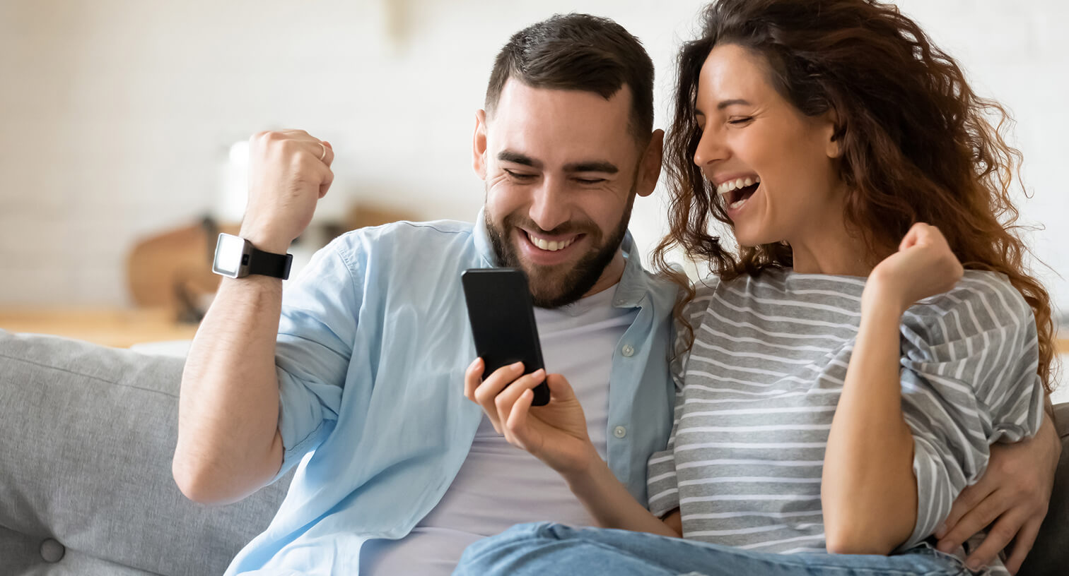 Pareja joven sonriendo sentada en un sofá mirando un celular.
